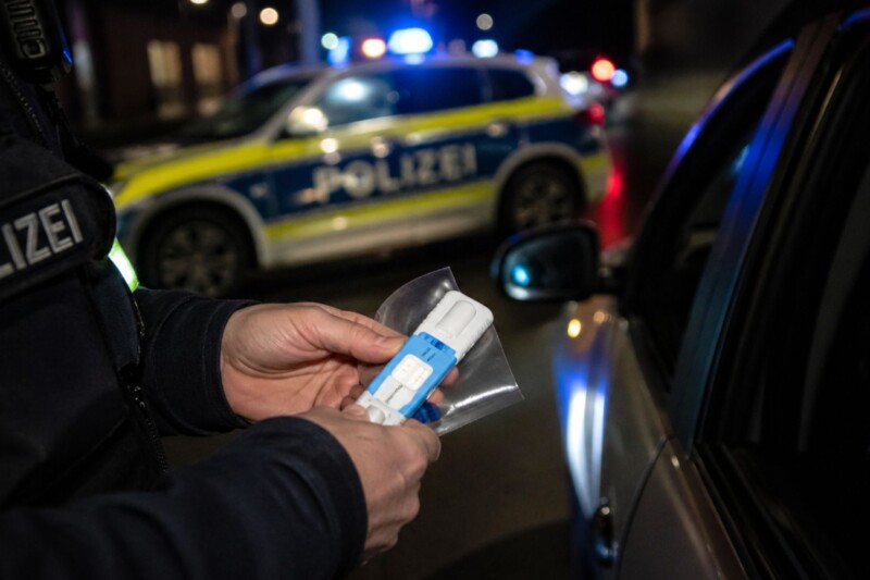 Ein Polizeibeamter hält einen Alkoholtest in der Hand, während ein Polizeiauto im Hintergrund steht.