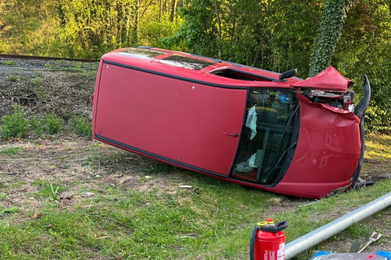 Ein umgekipptes rotes Auto liegt am Rand einer Bahnstrecke, umgeben von Grünpflanzen und einem Feuerlöscher.