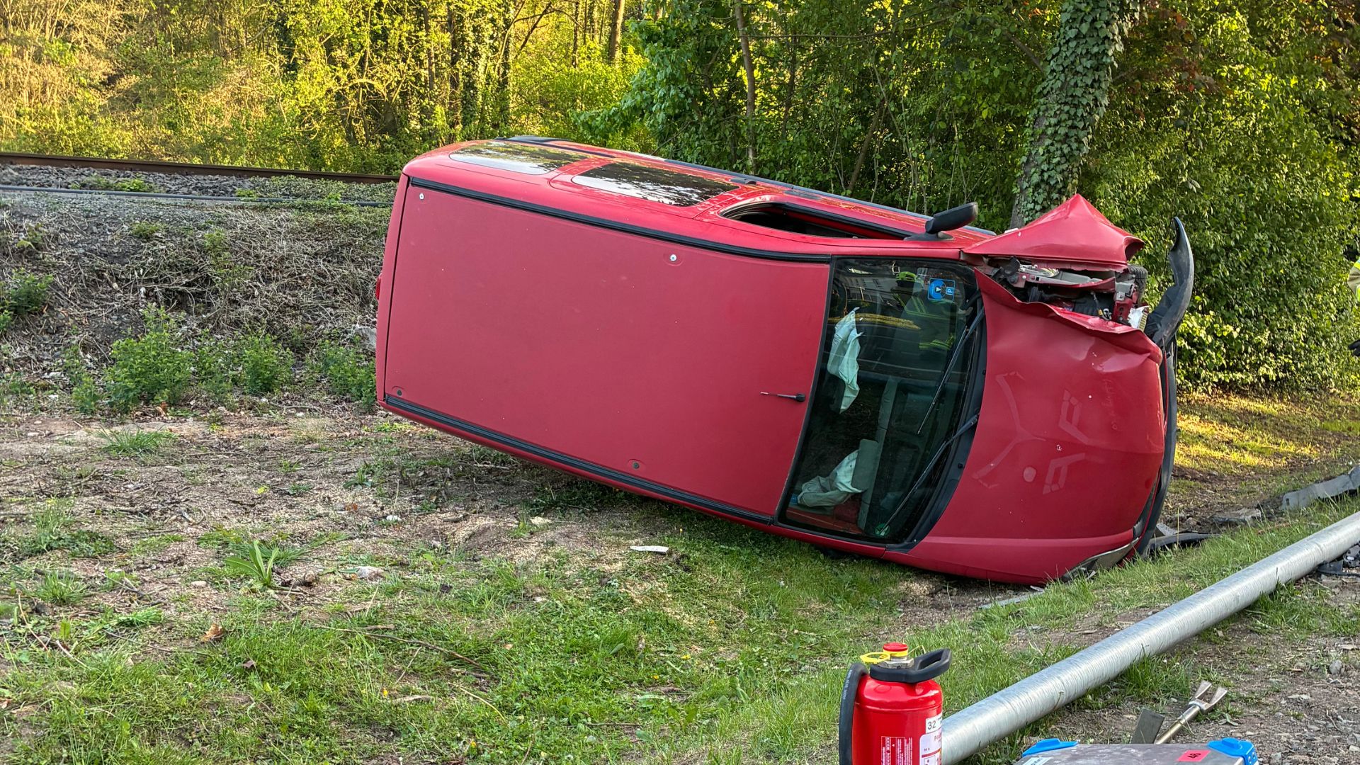 Ein umgekipptes rotes Auto liegt am Rand einer Bahnstrecke, umgeben von Grünpflanzen und einem Feuerlöscher.