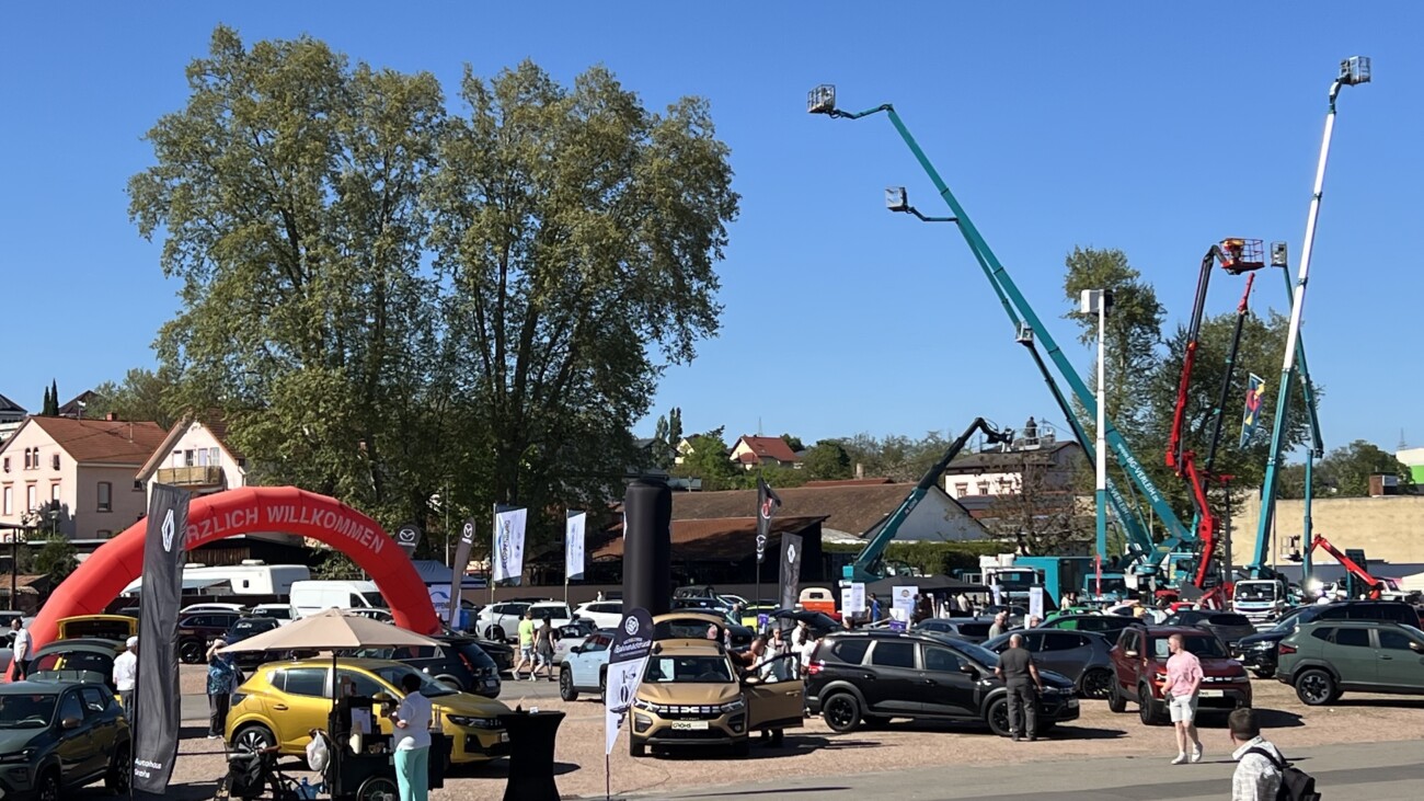 Ein Auto-Event mit verschiedenen Fahrzeugen und Hebebühnen unter blauem Himmel und Bäumen im Hintergrund.