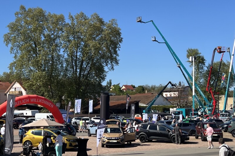 Ein Auto-Event mit verschiedenen Fahrzeugen und Hebebühnen unter blauem Himmel und Bäumen im Hintergrund.