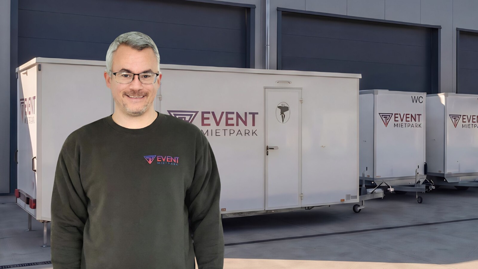 Ein Mitarbeiter von Pfannebecker Eventservice steht vor mobilen Toilettenanhängern in einer modernen Lagerhalle.