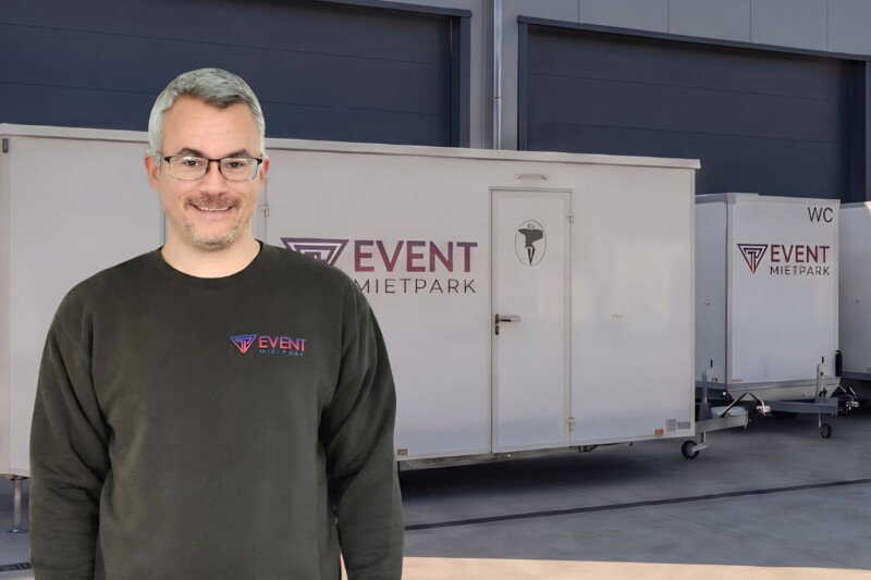 Ein Mitarbeiter von Pfannebecker Eventservice steht vor mobilen Toilettenanhängern in einer modernen Lagerhalle.