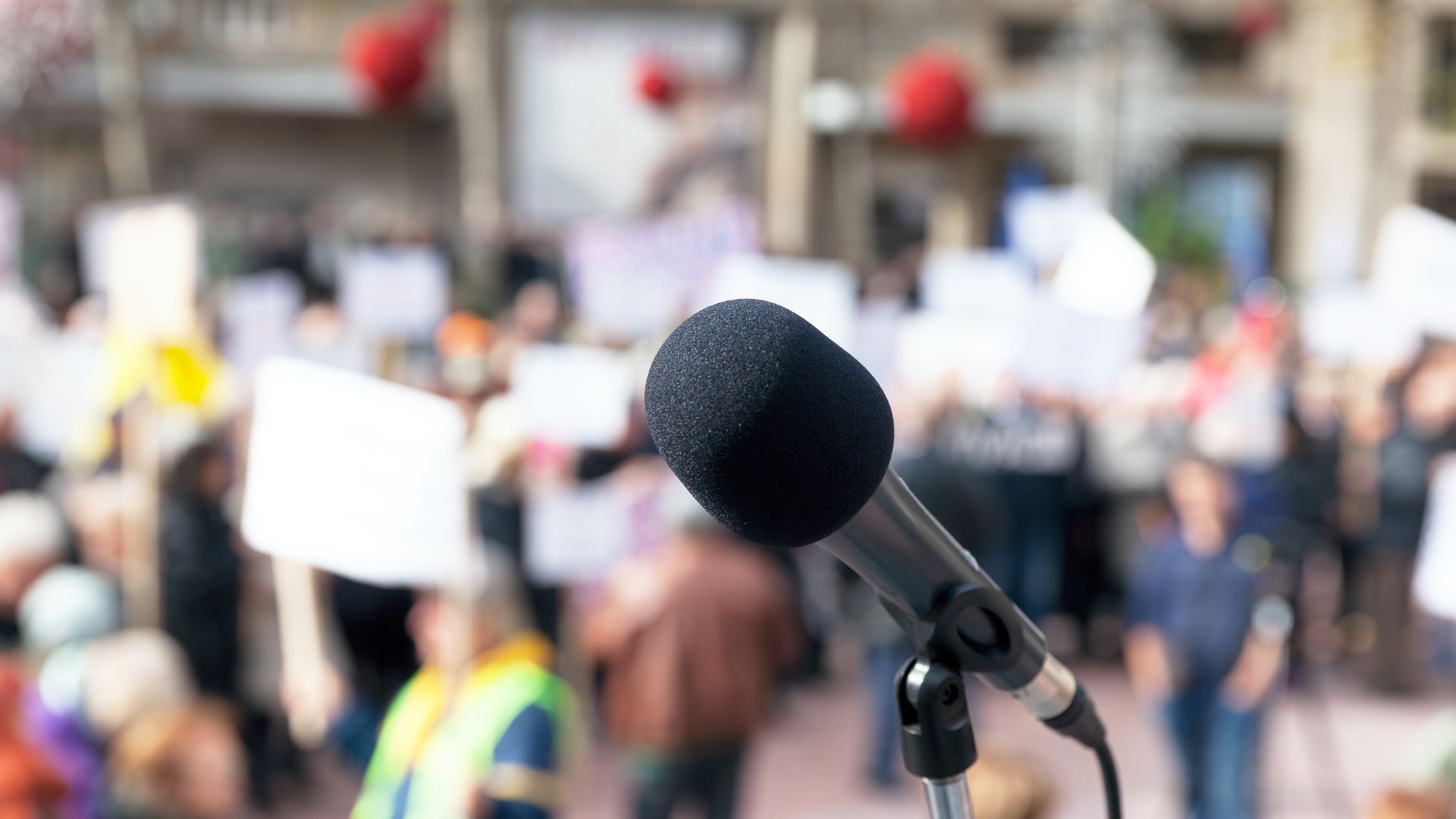 Mikrofon im Vordergrund, während eine Menschenmenge mit Plakaten im Hintergrund protestiert.