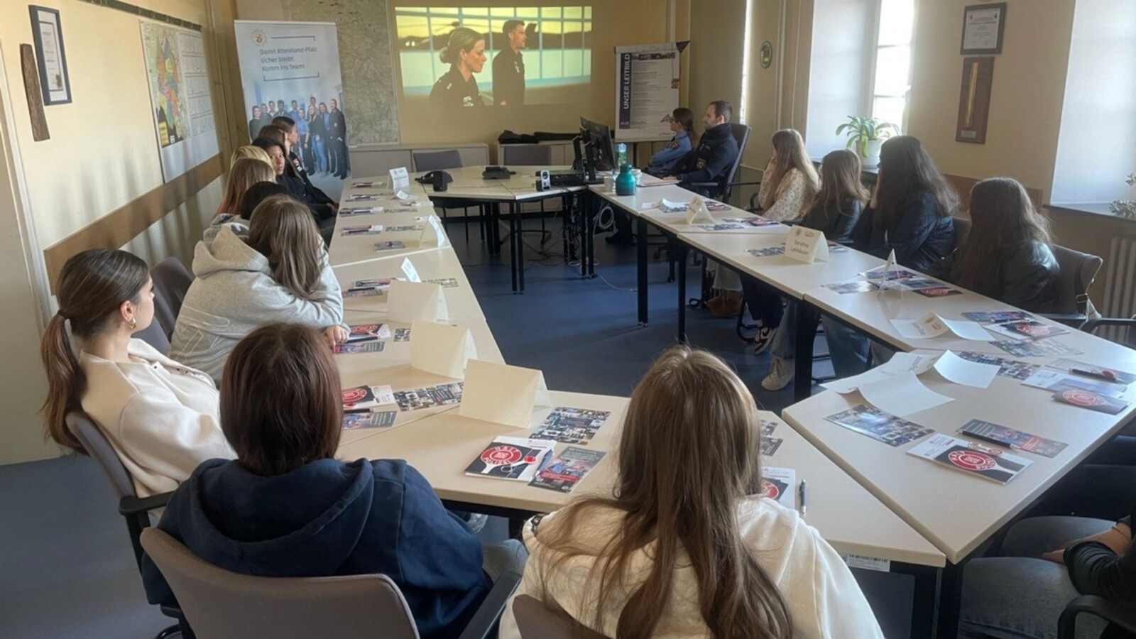 Eine Gruppe von Jugendlichen sitzt an einem Tisch und hört einem Vortrag zu, während sie Informationsmaterialien vor sich haben.