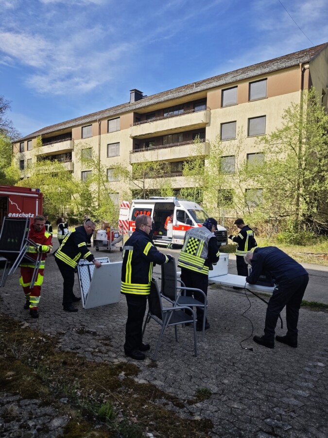 Rettungskräfte arbeiten an einem Einsatzort mit Möbeln und Rettungsfahrzeugen im Hintergrund.
