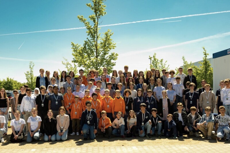 Gruppenfoto von Schülern und Lehrern im Freien, umgeben von Bäumen und einem blauen Himmel.