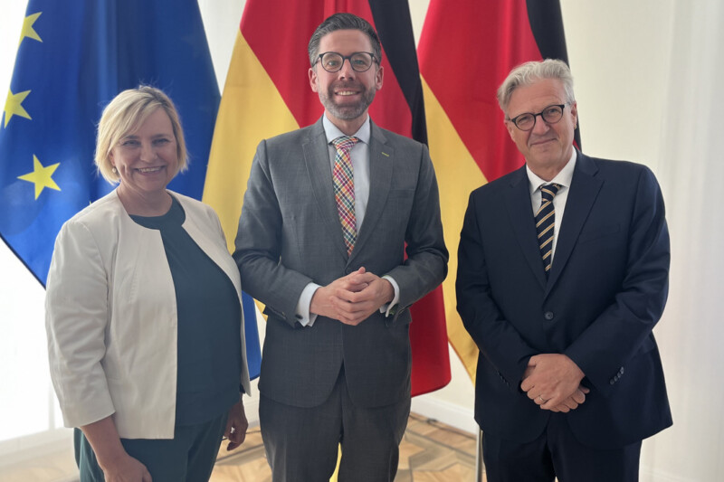 Drei Personen stehen vor den Flaggen der EU und Deutschlands, lächeln und posieren für ein Foto.