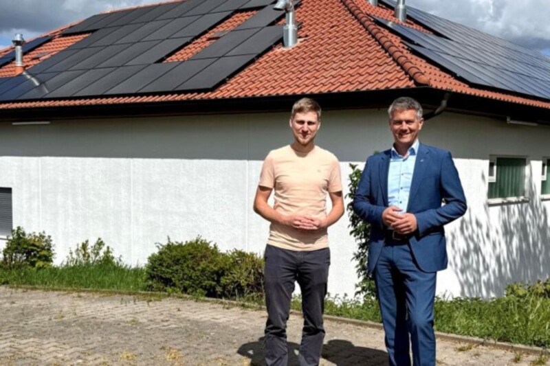 Zwei Männer stehen vor einem Haus mit Solarpanelen auf dem Dach, umgeben von Grünflächen.
