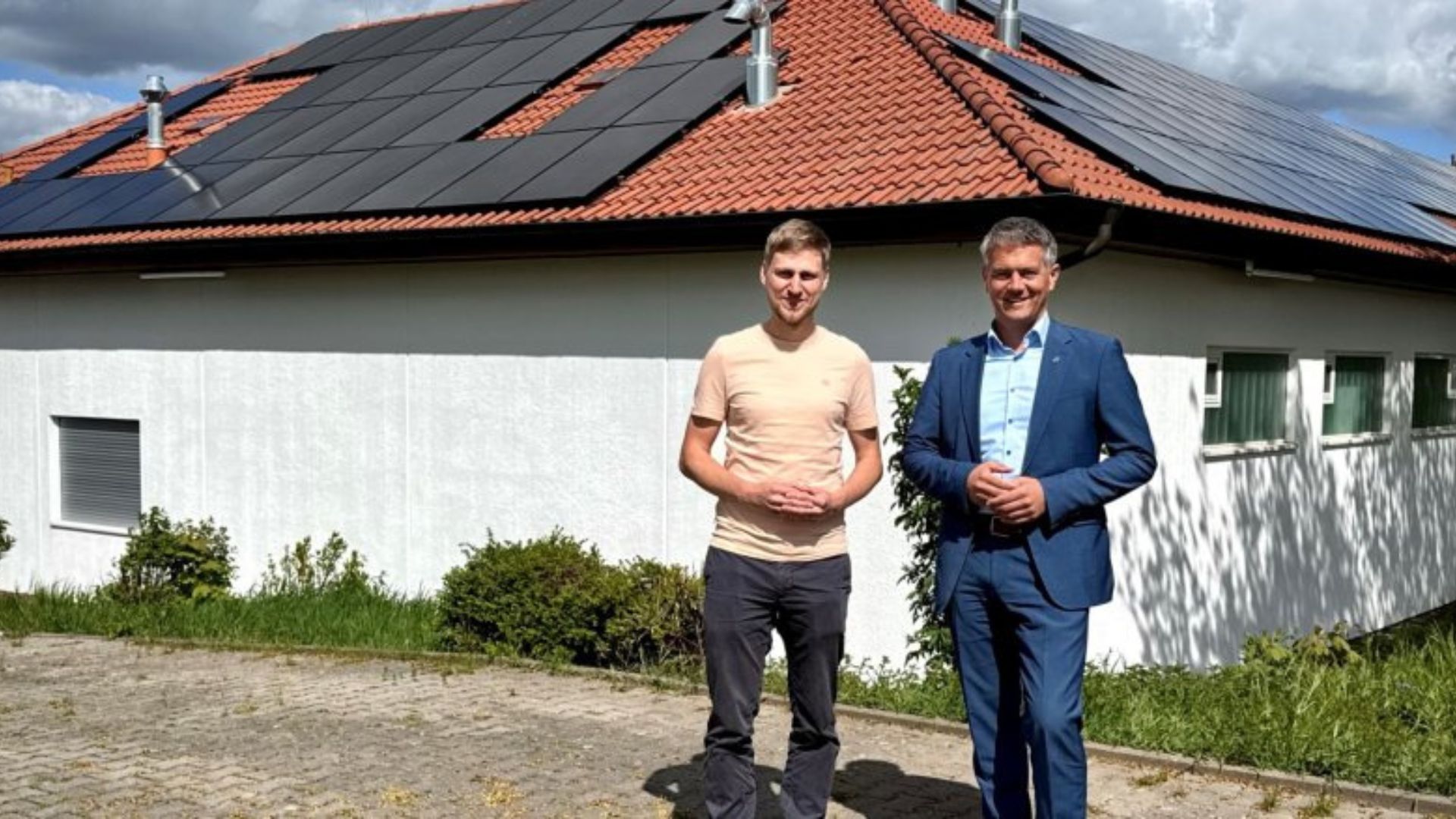 Zwei Männer stehen vor einem Haus mit Solarpanelen auf dem Dach, umgeben von Grünflächen.