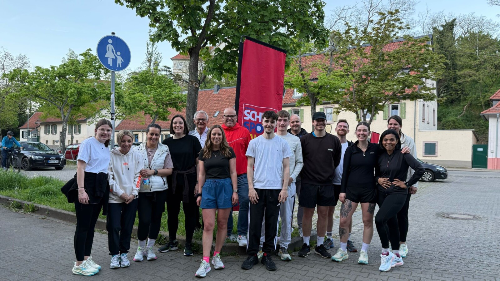 Gruppe von Menschen steht vor einem Banner, umgeben von Bäumen und Häusern, bei einem Sportevent im Freien.