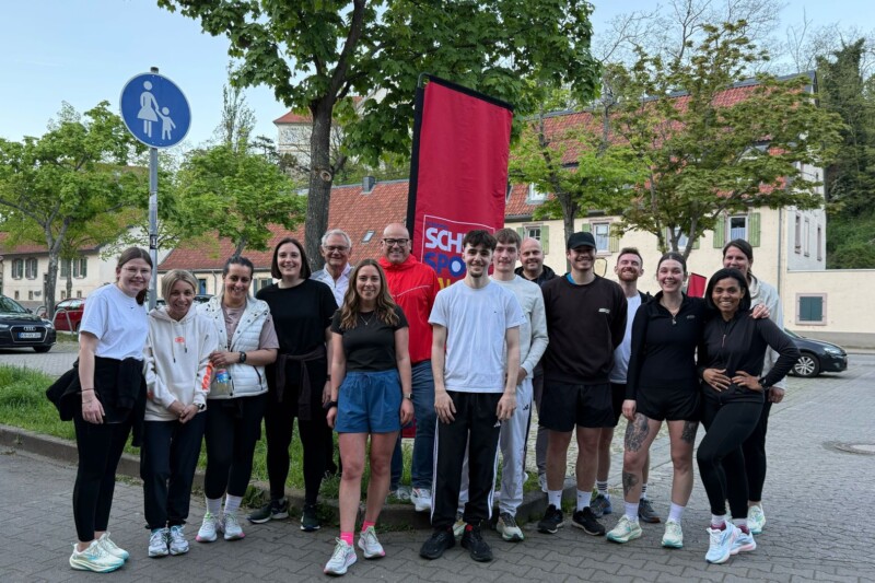 Gruppe von Menschen steht vor einem Banner, umgeben von Bäumen und Häusern, bei einem Sportevent im Freien.