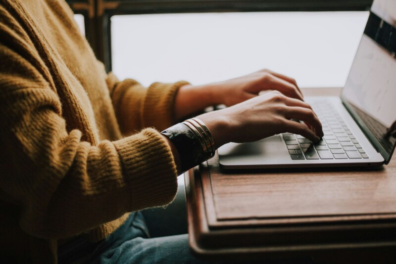 Eine Person tippt auf einem Laptop, während sie an einem Holztisch sitzt. Die Umgebung ist hell und einladend.