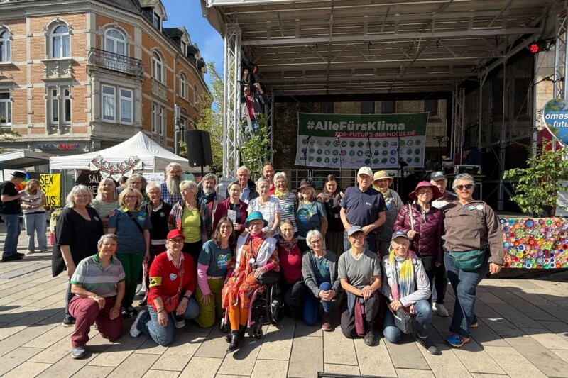 Eine Gruppe von Menschen steht auf einem Platz während einer Klima-Demonstration mit Schildern und einer Bühne im Hintergrund.