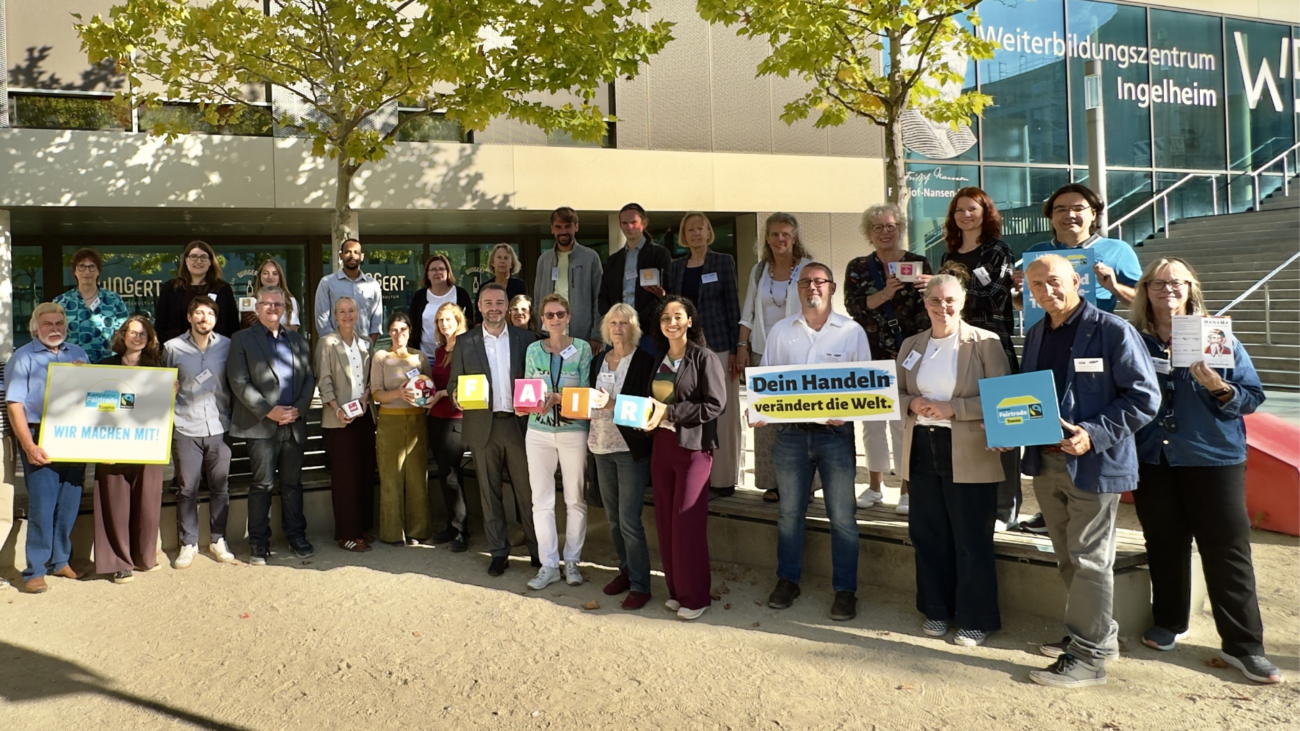 Zu sehen ist eine größere Gruppe von Menschen vor dem Weiterbildungszentrum Ingelheim. Einige Teilnehmer halten Schilder und Tafeln zum Thema Fairtrade und fairer Handel in den Händen. Die Gruppe steht im Außenbereich vor dem Gebäude, umgeben von Bäumen und Grünflächen.