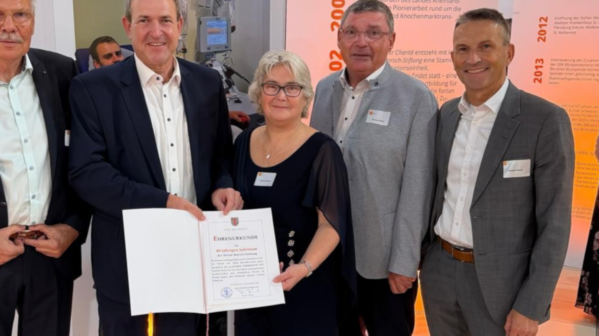 Zu sehen sind fünf Personen, die nebeneinander vor einer Ausstellung zur Geschichte der Stefan-Morsch-Stiftung stehen. Zwei Personen in der Mitte halten gemeinsam eine geöffnete Ehrenurkunde zum 40-jährigen Jubiläum der Stiftung. Im Hintergrund sind Ausstellungstafeln mit Jahreszahlen, Texten und Bildern zur Entwicklung der Stiftung zu erkennen.