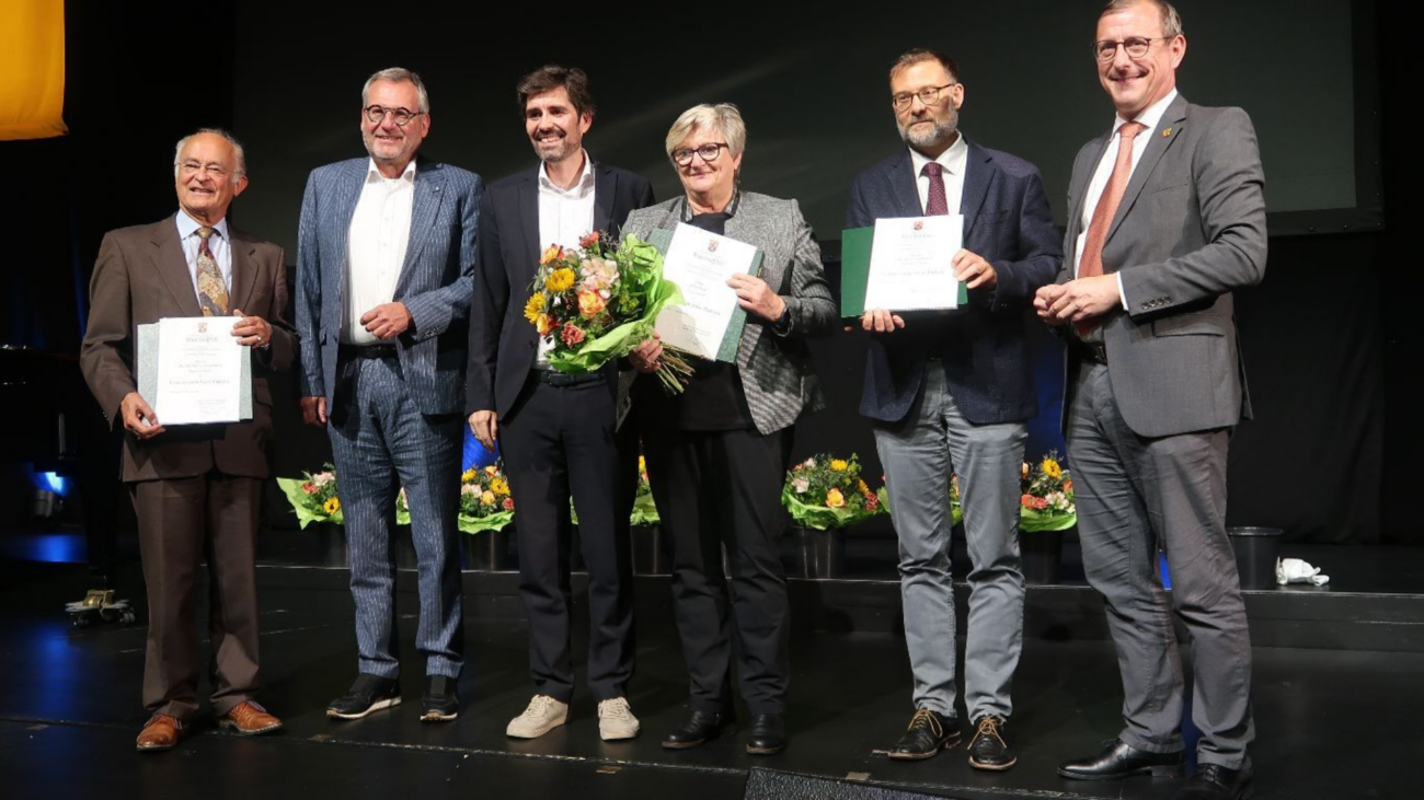 Zu sehen sind sechs Personen auf einer Bühne bei einer Auszeichnungsveranstaltung. Mehrere von ihnen halten Urkunden, eine Person zusätzlich einen Blumenstrauß. Im Hintergrund ist eine dunkle Bühnenwand zu sehen.