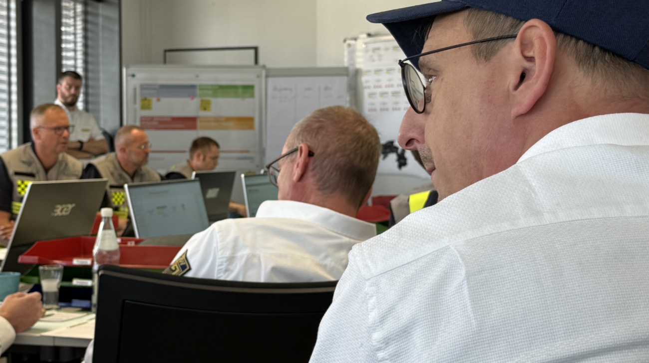 Zu sehen sind mehrere Personen in einem Raum, die an Tischen mit Laptops und Unterlagen arbeiten. Im Vordergrund stehen zwei Männer, während im Hintergrund weitere Teilnehmer vor Bildschirmen sitzen. An den Wänden hängen Übersichten und Lagekarten, wodurch die Szene wie eine Besprechung oder Einsatzkoordination wirkt.