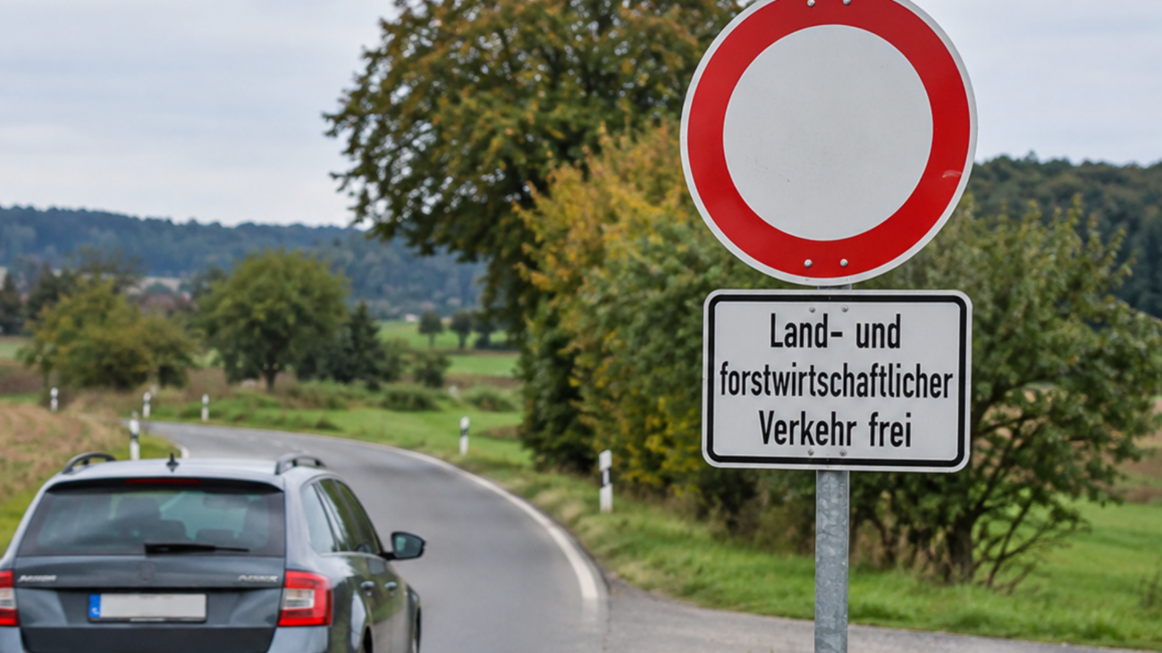 Zu sehen ist eine ländliche Straße mit einem Auto, das an einem Verkehrszeichen vorbeifährt. Das Schild zeigt ein Durchfahrtsverbot mit dem Zusatz „Land- und forstwirtschaftlicher Verkehr frei“. Im Hintergrund sind Felder, Bäume und einzelne Häuser zu erkennen.